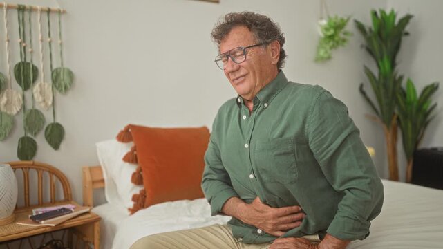 Man clutching stomach while seated on bed in a residential building, hand pressed to abdomen and wincing with visible stomachache; pain vulnerability.