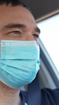 A man wearing a blue mask is seen in the car. His expression suggests attentiveness as he looks outside the window while driving.