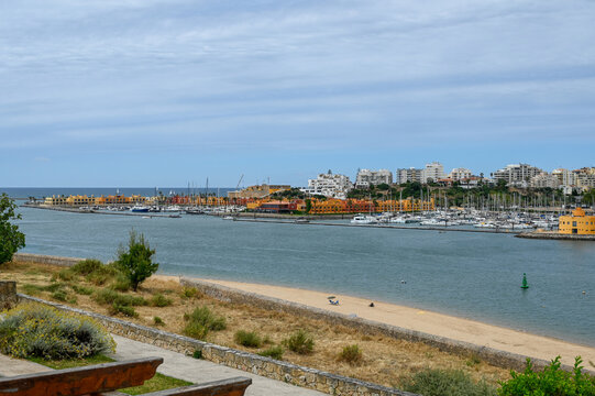 View of a marina with numerous sailboats and yachts docked, colorful buildings along the waterfront, a sandy beach in the foreground, and a calm sea in Algarvia, Azores, Portugal.