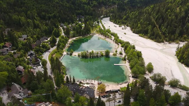 Aerial view of Lake Jasna in Kranjska Gora, Slovenia, with turquoise alpine ponds, forested slopes, village homes and pale riverbed beneath Vr&scaron;ič Pass.