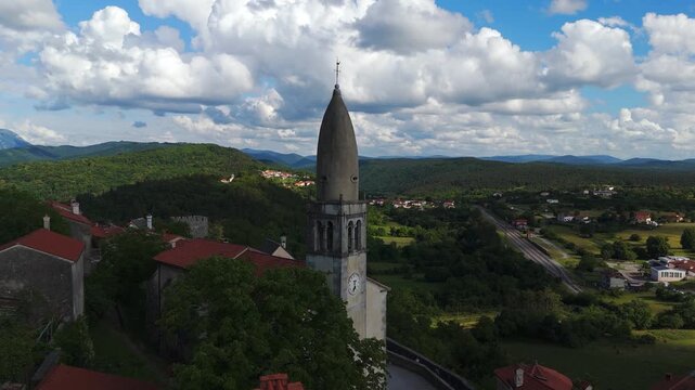 Tilting aerial view, St. Daniel Church bell tower in &Scaron;tanjel, Slovenia, overlooking the Karst countryside with rolling hills, traditional architecture and summer clouds