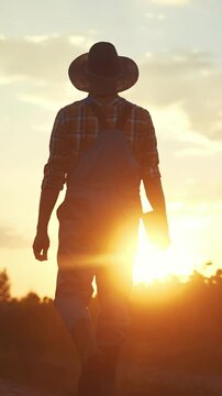 A man stands silhouetted against the sunset. The man is wearing a hat and overalls, observing the tranquil scene of the evening sun setting.