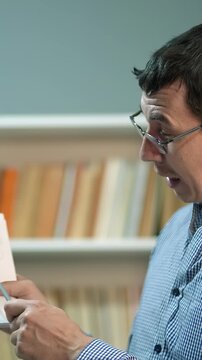 A man in glasses reads a book intently, surrounded by shelves filled with books. The setting features a calm atmosphere with a focus on the man and the books.