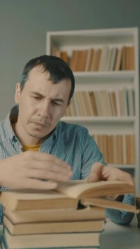 A man reads a book while seated at a table. Books are stacked around him, and a shelf filled with books is visible in the background.