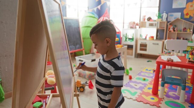 Little boy draws on easel and points finger to scribble in colorful classroom filled with toys and foam number mat; curiosity.
