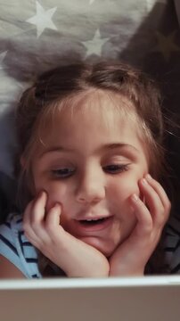 A young girl is happily watching something on a screen. Her expression shows interest and joy as she rests her chin on her hands while watching the screen.