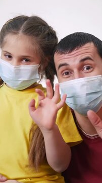 A man and a girl wear masks while smiling. The girl is in a yellow shirt and the man waves his hand, both appearing cheerful in a domestic setting.