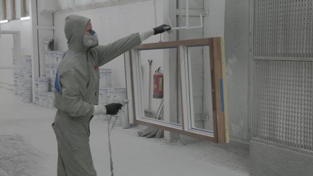 Worker in protective suit spray painting a wood frame window inside an industrial factory booth with paint containers and ventilation wall
