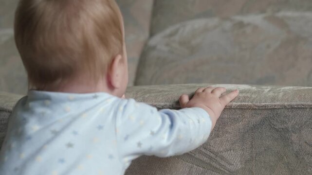 Infant steadies with furniture support. Young child tries to balance while holding onto furniture. Baby in soft clothing works to maintain upright posture using nearby furniture in warm room