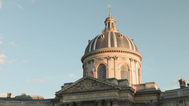 Close-up shot of the majestic dome of the Institut de France in Paris, illuminated by warm golden hour sunlight against a soft blue sky with a French flag waving gently.