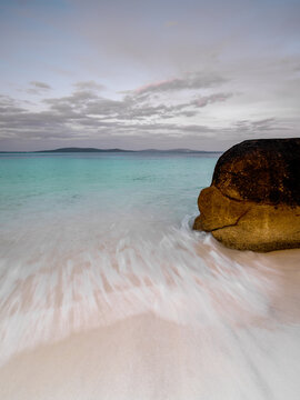View of Whalers Cove with turquoise water, a large granite boulder on the sandy shore, and a soft cloudy sky at sunset in Albany, WA, Australia.