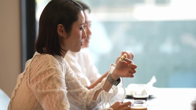 Soft daylight illuminates the scene. Woman, wearing a light-colored knit top, sits at a table. She holds a small snack, gesturing with her other hand