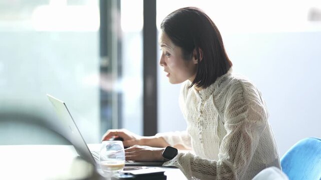 Focused woman types on laptop at desk. Soft light streams through large windows. Colleague stands nearby, gesturing subtly. Professional setting, calm concentration. Great for business, teamwork
