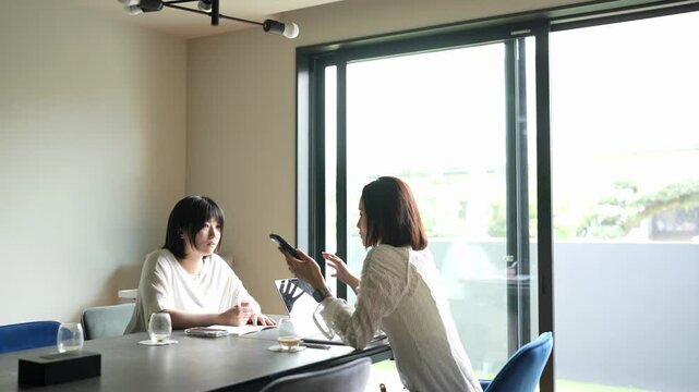 Two women sit at a modern table. One uses a phone, the other looks at a laptop. Natural light streams through floor-to-ceiling windows. They appear focused, yet relaxed in a professional setting