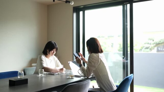 Two women collaborate in a bright, modern office space. One focuses on documents, the other checks her phone. Natural light streams through floor-to-ceiling windows