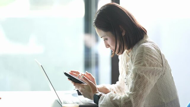 Woman focuses intently on her smartphone. She sits beside an open laptop. Natural light streams through large windows. She wears a soft, textured blouse. Her posture suggests concentration