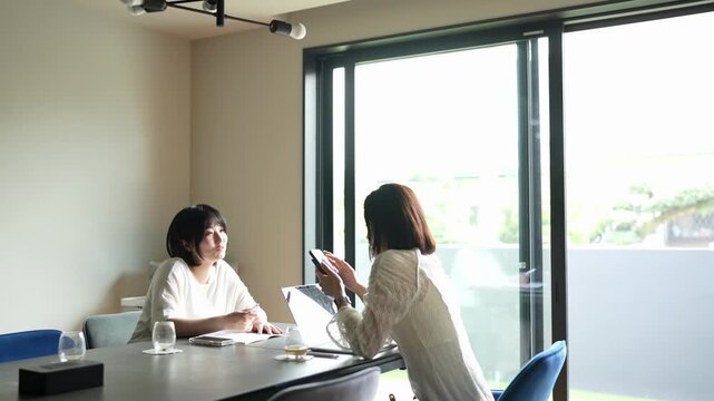 Two women sit at a table in a bright room. One reads while the other holds a phone. They appear calm and focused on their tasks. Natural light fills the space through large windows