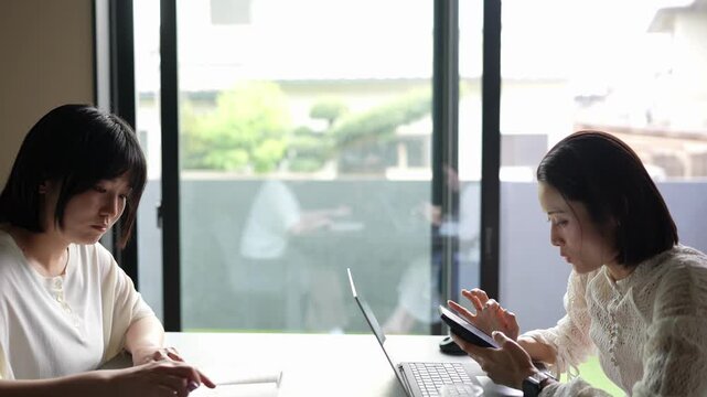 Two women work side by side in a bright, modern office. One uses a laptop, the other holds a tablet, focused on tasks. Natural light floods the space through large windows