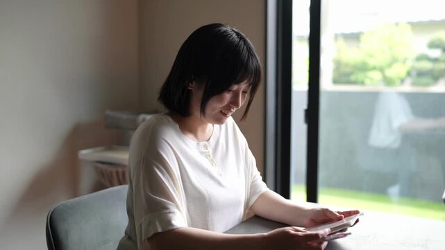 Soft light glows through large glass window. Woman sits quietly, focused on tablet device. Her dark hair frames calm, concentrated expression. Peaceful home setting, gentle focus, quiet moments