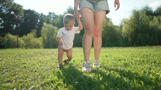 Happy Toddler takes first step on grass with mom. Mom guides toddler on grassy park path. Happy Toddler learning to walk on grass with mom support. Happy moment as toddler steps on grass with mom.