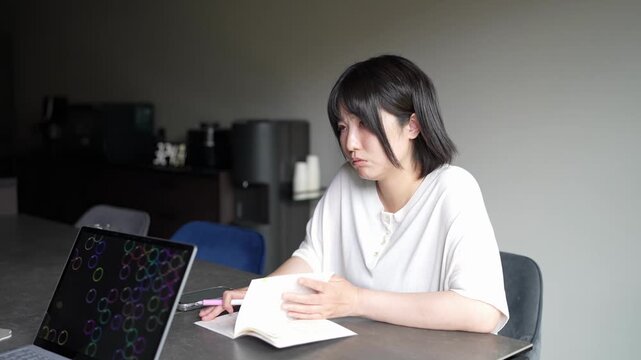 Young woman focuses on notebook beside laptop. She writes, eyes slightly downcast, pensively engaged. Laptop screen glows with colorful circle pattern. Quiet, focused atmosphere, study or work session