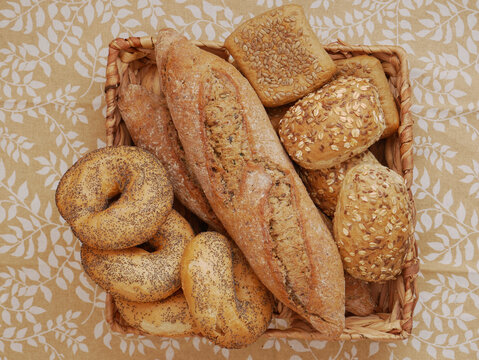 A basket is filled with various types of bread, including a baguette, bagels, and rolls. The breads are arranged on a tablecloth with a leaf pattern.