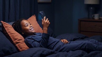 Fototapeta premium Happy young African American girl lying in bed at night using a smartphone with a glowing screen
