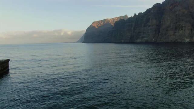 Aerial perspective slowly revealing the impressive Los Gigantes cliff formations at sunset. Calm Atlantic Ocean waters and a concrete pier