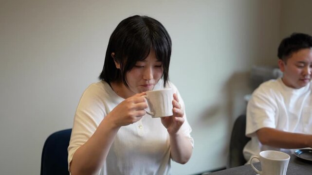 Soft light fills calm, cozy room. Woman sips warm drink, smiling gently. Man sits beside her, focused on screen. Both dressed in plain white shirts. Simple, quiet moment, warm and friendly