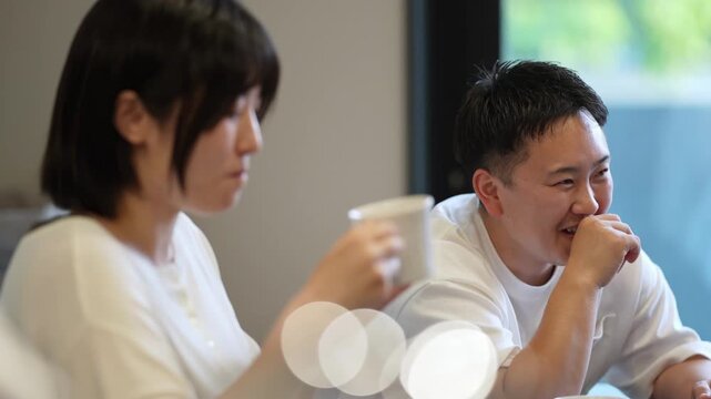 Two people in white shirts sit at a table. They share a quiet, relaxed moment together. One sips from a cup while the other smiles gently. Soft light and blurred bokeh enhance the calm