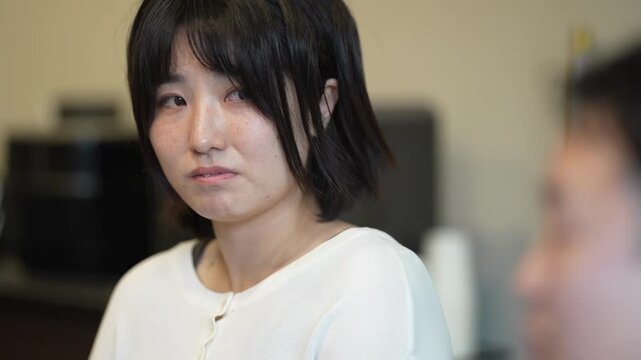 Young woman wears white top, gazes directly. She shows mild concern, slightly frowning, in softly lit room. Blurred figure sits beside her, focused on conversation