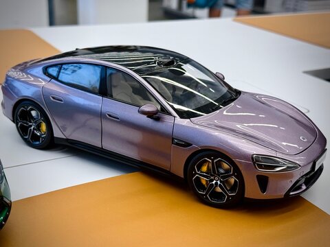 Xiaomi SU7. Die-cast scale model car displayed indoors, possibly in a Xiaomi store. The model is a sleek, modern sedan in light purple with a glossy finish. Features include realistic detailing