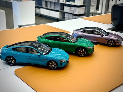 Three scale die-cast model cars are displayed on a table in a Xiaomi store. The models include a blue, green, and purple car, each with sleek, modern designs and detailed features like tinted windows 