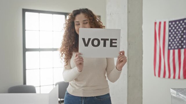 Woman, young hispanic with curly hair, smiles and holds vote sign with hands in polling building by an american flag; civic pride.
