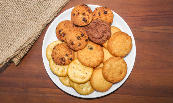 Assorted cookies and biscuits on a white plate on a wooden table. A collection of sweet baked snacks including chocolate cookies, butter biscuits, crackers, and sandwich cookies on a plate.