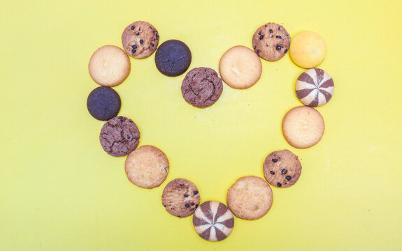 Assorted cookies and biscuits arranged in love shape isolated on Yellow background. Collection of sweet baked snacks including chocolate cookies, butter biscuits, crackers and sandwich cookies.