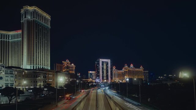 Timelapse of vehicle light trails flowing along a busy avenue at night, with a soft glow of distant buildings in the background.