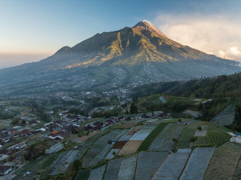 Aerial view of the majestic Mount Merapi volcano rising above the village with terraced agricultural fields and lush green slopes in Selo, Central Java, Indonesia.