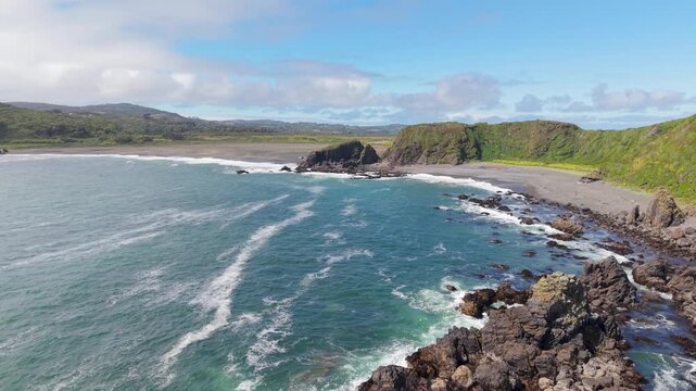 Cinematic landscape aerial view of a drone flying forward towards a secluded sandy beach and lush green hills on the rugged Pacific coastline of Chilo&eacute; Island in Chilean Patagonia.