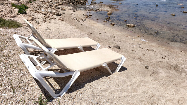 Two empty sun loungers on a sandy and rocky beach in Protaras, Cyprus. A place for summer relaxation near the water, seaside relaxation, and tourism and travel concepts.