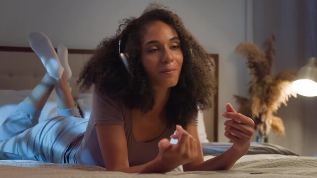 Happy African American woman using phone listening music in headphones lying on bed in cozy bedroom joyous female girl sing listen sound in earphones browsing mobile app singing joy at night insomnia
