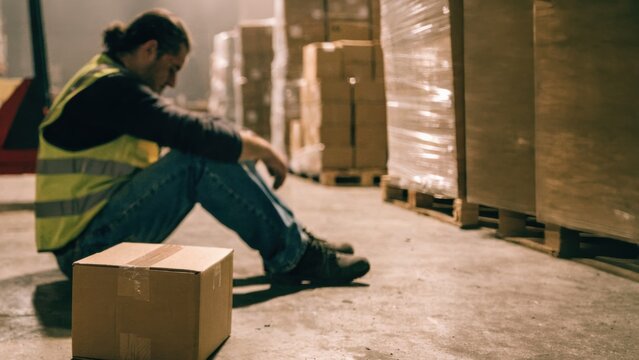 A man in a safety vest sits on the floor of a warehouse, leaning on a box, appearing tired or stressed, surrounded by stacked pallets and boxes.