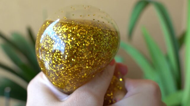 Close-up video of a sparkling golden squishy toy with a cute cartoon face slowly compressed in hand. Concept of emotional regulation, relaxation, and stress relief