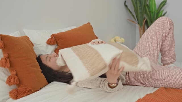 Young chinese woman reclining on bed, reaching hand upward while holding decorative tassel pillow among orange cushions and throw in a bright bedroom; serenity.