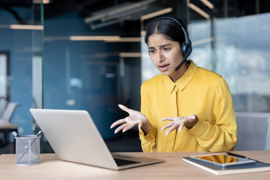 Upset and worried young Indian businesswoman in headset talking emotionally in office on laptop camera, gesturing with hands