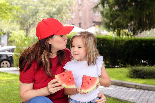 Mother kissing her daughter while holding watermelon slices in a sunny park. Loving family bond, healthy summer snack, outdoor lifestyle, childhood care, and affection concept