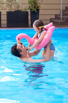 Happy father lifting his little daughter on a pink flamingo float in a swimming pool. Family bonding, summer fun, water safety, active parenting, and vacation lifestyle concept