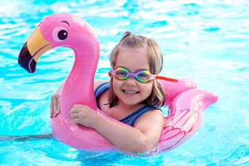Smiling little girl wearing colorful swimming goggles floating on a pink flamingo inflatable in a blue pool. Summer fun, water safety, childhood happiness, and vacation concept © AMR Studio
