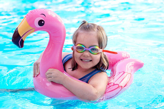 Smiling little girl wearing colorful swimming goggles floating on a pink flamingo inflatable in a blue pool. Summer fun, water safety, childhood happiness, and vacation concept