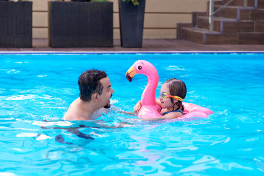 Father teaching his little daughter to swim in a pool with a pink flamingo float. Happy family bonding, water safety, summer vacation, and active parenting concept
